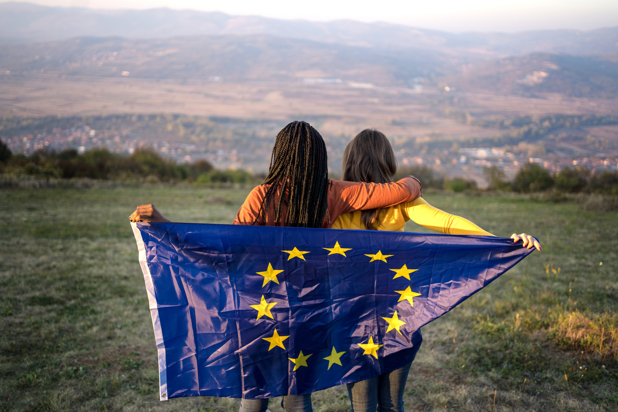 Two young people on a hill looking into the horizon holding a European flag behind their backs